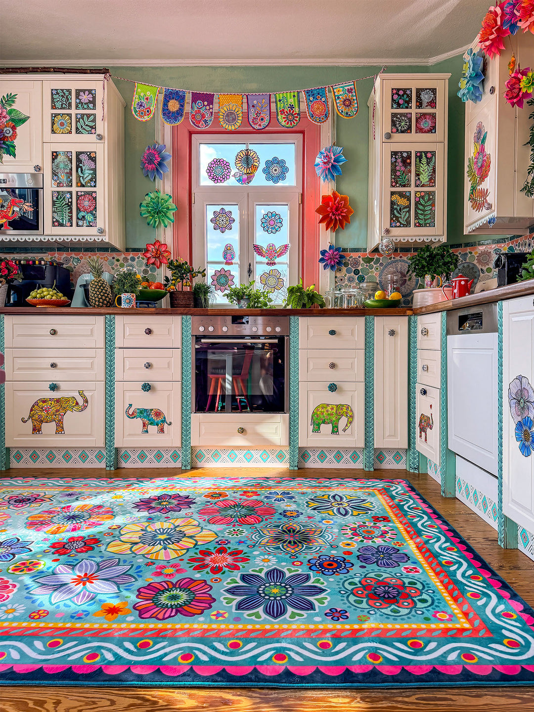 Großer Teppich in Türkis mit farbenfrohen Blumen- und Mandala-Mustern in Pink, Gelb, Rot, Blau, Grün und Lila liegt auf Holzboden in einer kreativ gestalteten Küche. Im Hintergrund stehen helle Küchenschränke mit dekorativen Elefantenmotiven, Holzarbeitsplatten, Pflanzen, Obstschalen und bunte Fensterdekorationen. Der florale Teppich bringt lebendige Farben, gemütliche Wärme und verspielten Boho-Stil in den Küchenbereich.