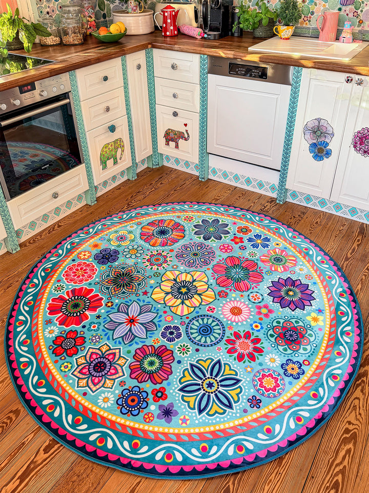 Runder Teppich in Türkis mit farbenfrohen Blumen- und Mandala-Mustern in Pink, Gelb, Rot, Blau, Grün und Lila liegt auf warmem Holzboden in einer kreativ gestalteten Küche. Im Hintergrund stehen helle Küchenschränke mit dekorativen Elefanten- und Blumenmotiven, Holzarbeitsplatten, Pflanzen sowie moderne Küchengeräte. Der bunte Teppich setzt einen lebendigen Mittelpunkt und bringt gemütlichen Boho-Stil in den Küchenbereich.