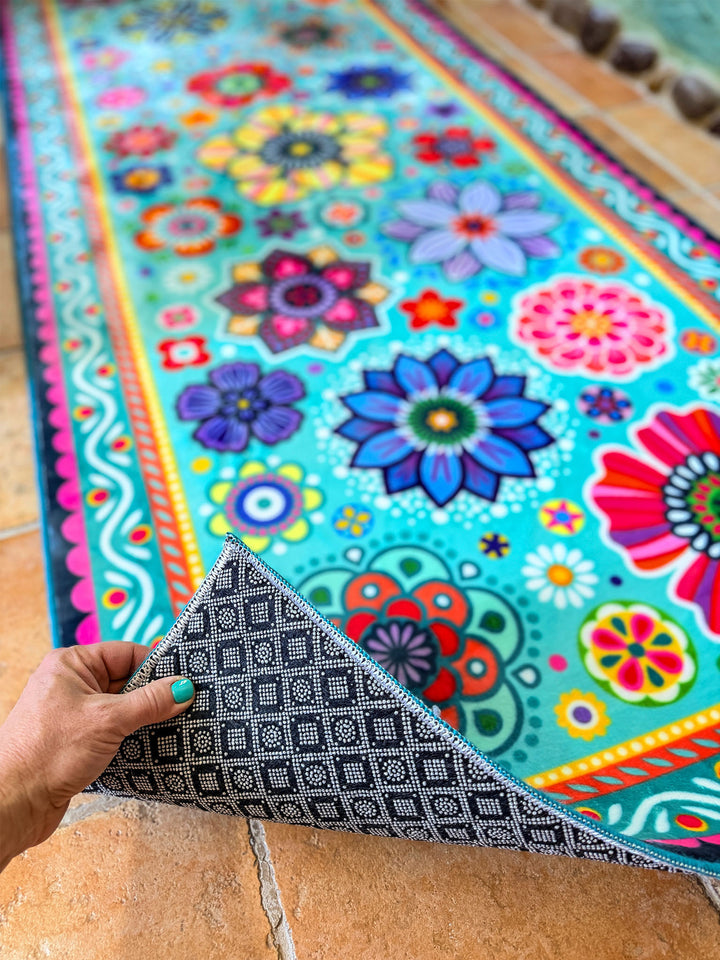 Schmaler rechteckiger Badteppich in Türkis mit farbenfrohen Blumen- und Mandalamotiven in Pink, Blau, Gelb, Rot und Grün liegt auf terrakottafarbenen Fliesen vor einer Badewanne. Das kreative Badezimmer ist mit kunstvollen Mosaikwänden, Naturstein-Details und Pflanzen dekoriert. Große grüne Blätter und ein Fenster mit Tageslicht schaffen eine frische, natürliche Atmosphäre. Der lebendige Teppich setzt einen fröhlichen Farbakzent im individuell gestalteten Boho-Bad.