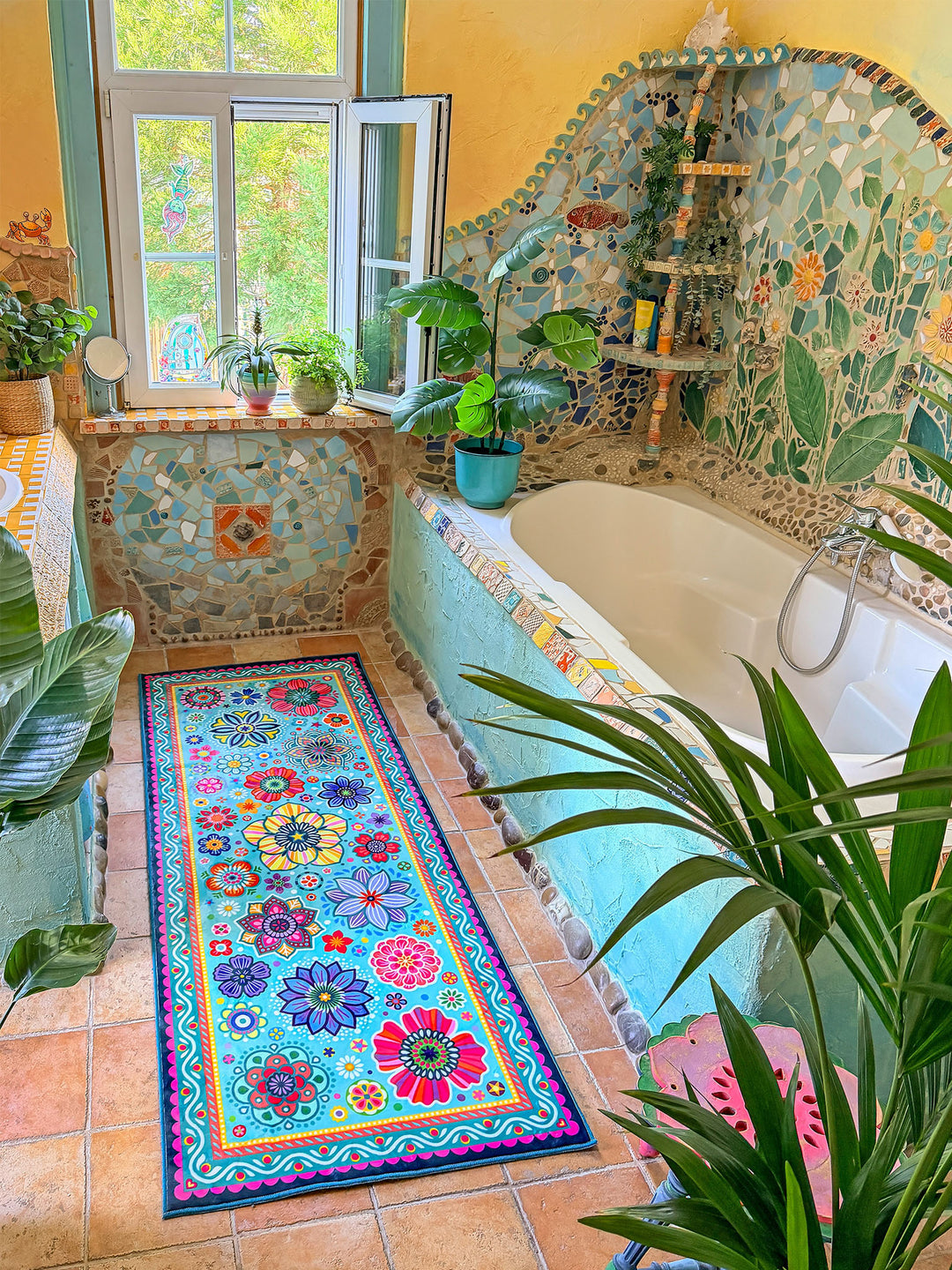 Schmaler rechteckiger Badteppich in Türkis mit farbenfrohen Blumen- und Mandalamotiven in Pink, Blau, Gelb, Rot und Grün liegt auf terrakottafarbenen Fliesen vor einer Badewanne. Das kreative Badezimmer ist mit kunstvollen Mosaikwänden, Naturstein-Details und Pflanzen dekoriert. Große grüne Blätter und ein Fenster mit Tageslicht schaffen eine frische, natürliche Atmosphäre. Der lebendige Teppich setzt einen fröhlichen Farbakzent im individuell gestalteten Boho-Bad.