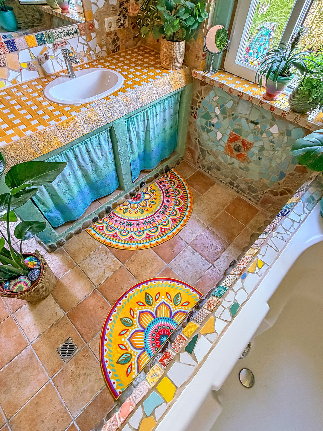 Kreativ gestaltetes Badezimmer im Boho-Stil mit zwei halbrunden Badteppichen in leuchtendem Gelb. Die Matten zeigen detailreiche Mandala- und Blumenmuster in Pink, Türkis, Blau, Orange und Grün und liegen auf warmen Natursteinfliesen vor Waschtisch und Badewanne. Der Raum ist mit Mosaikwänden, dekorativen Fliesen, Pflanzen und türkisfarbenen Stoffvorhängen unter dem Waschbecken gestaltet. Tageslicht fällt durch das Fenster und sorgt für eine freundliche, mediterrane Wohlfühlatmosphäre.