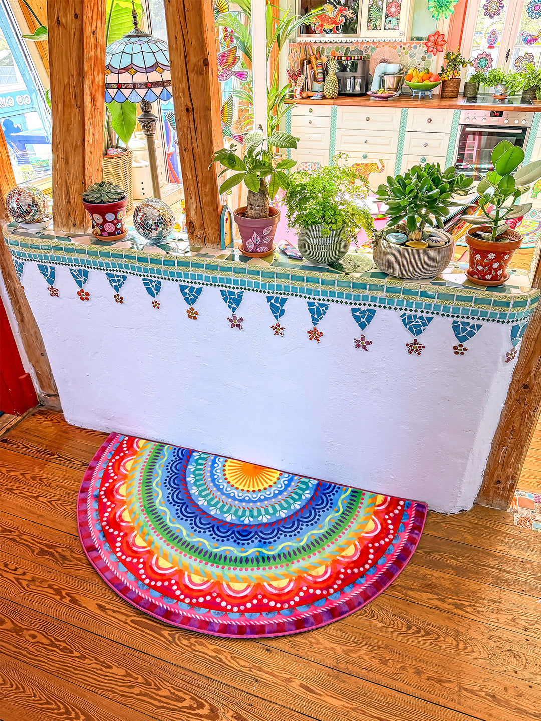 Halbrunder Teppich mit farbenfrohem Mandala-Muster in Blau, Türkis, Gelb, Rot, Pink und Lila liegt auf warmem Holzboden vor einer offenen Wohnküche. Auf der gemauerten Fensterbank stehen zahlreiche Zimmerpflanzen in dekorativen Töpfen. Im Hintergrund sind eine helle Küche, Holzdetails und bunte Dekorationen zu sehen. Die Szene wirkt lebendig, kreativ und gemütlich mit fröhlichem Boho-Wohnstil.
